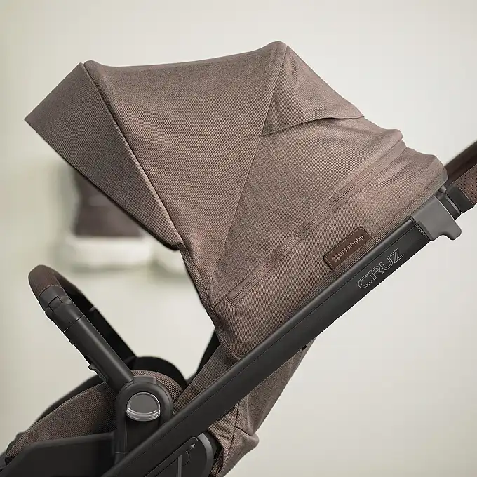 Side profile of a UPPAbaby Cruz stroller in brown fabric, showing the canopy, leather handle, and "Cruz" branding on the frame.